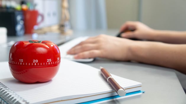 A tomato kitchen timer on a desk next to a person using a keyboard to type on a computer